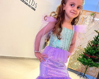 May include: A young girl wearing a purple and turquoise mermaid costume with a sequined bodice and tulle skirt. She is wearing a flower crown and white shoes. The costume is from the brand "IKRAKIDS PERSONALIZED COSTUMES AND DRESSES".