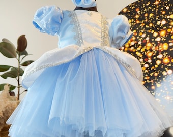 May include: A light blue princess dress with a matching bow headband. The dress features a white bodice with silver trim and puffy sleeves. The skirt is layered with tulle. The dress is displayed on a black mannequin.