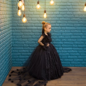 May include: A young girl wearing a black tulle dress with a long train and a sequined bodice. She is standing in front of a blue brick wall with string lights hanging from the ceiling.