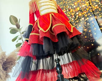 May include: A red and black costume with gold accents, displayed on a black mannequin. The costume features a military-style jacket with gold fringe epaulets and a tiered skirt with layers of red and black tulle. The costume is designed for a performance.