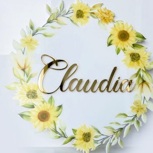 Può includere: Un cartello circolare bianco con una ghirlanda floreale dorata e il nome "Claudia" scritto in lettere dorate.