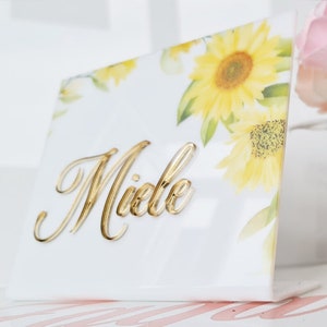 Può includere: Un cartello in acrilico bianco con un design a girasole e il nome "Miele" scritto in lettere dorate.