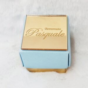 Può includere: Una piccola scatola regalo quadrata con un coperchio dorato e una base azzurra. Il coperchio è inciso con le parole "Benvenuto Pasquale" in una scrittura elegante. La scatola è su una superficie bianca e soffice.