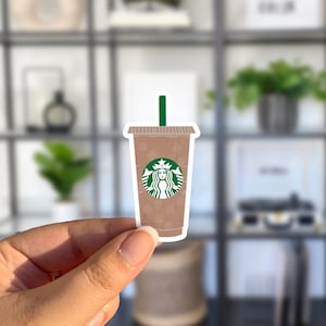 May include: A brown and white iced coffee cup with a green Starbucks logo and a green straw. The cup is being held by a hand.