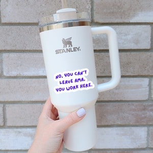 K&ouml;nnte beinhalten: Wei&szlig;e Edelstahl-Reisebecher mit schwarzem Griff und einem schwarz-wei&szlig;en Logo mit der Aufschrift "STANLEY". Der Becher hat einen Aufkleber mit der Aufschrift "NO, YOU CAN'T LEAVE AMA. YOU WORK HERE."