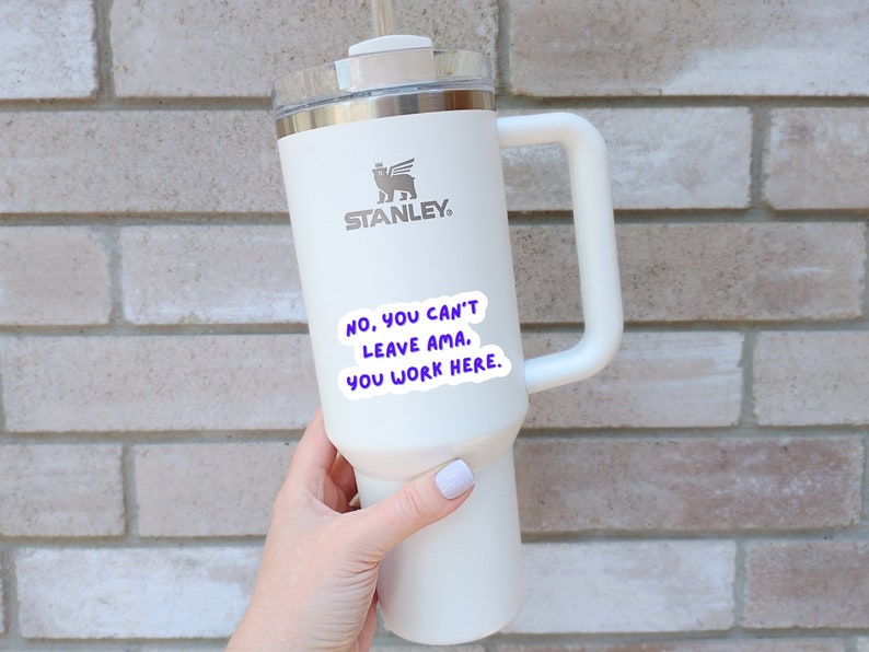 K&ouml;nnte beinhalten: Wei&szlig;e Edelstahl-Reisebecher mit schwarzem Griff und einem schwarz-wei&szlig;en Logo mit der Aufschrift "STANLEY". Der Becher hat einen Aufkleber mit der Aufschrift "NO, YOU CAN'T LEAVE AMA. YOU WORK HERE."