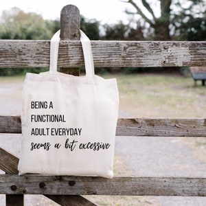 May include: A white canvas tote bag with black text that reads "Being a functional adult everyday seems a bit excessive". The bag is hanging on a wooden fence.