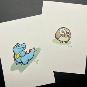 May include: Two hand-drawn illustrations on white paper. One features a blue cartoon crocodile with a yellow chest and red spikes. The other depicts a brown owl with green leaves. Both are on a green background.