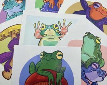 May include: A collection of art prints featuring colorful cartoon frogs in various poses. The frogs are depicted in shades of green, orange, and purple, with whimsical expressions. Some are perched on mushrooms or in front of colored circles.