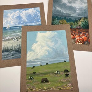 May include: Three framed landscape paintings. One depicts a beach scene with a sailboat and seagulls. Another shows a field of pumpkins with a carved jack-o'-lantern. The third features cows grazing on a green hillside under a cloudy sky.