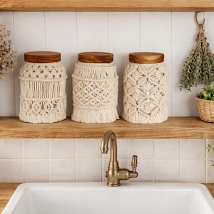 Peut inclure: Trois bocaux en macramé beige avec des couvercles en bois sur une étagère en bois. Un vase blanc avec des fleurs séchées est à gauche. Un robinet en laiton et un évier blanc sont au premier plan. Des paniers en osier avec des herbes et des plantes sont à droite.