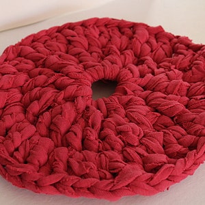 May include: A close-up of a red crocheted round rug with a hole in the center. The rug is made of thick, twisted yarn and has a textured surface.