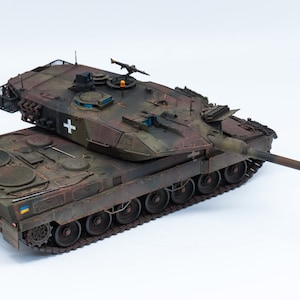 Puede incluir: Un modelo detallado de un tanque militar con un esquema de pintura de camuflaje. El tanque tiene un cañón grande y una ametralladora más pequeña en la parte superior. El tanque está pintado con un patrón de camuflaje verde, marrón y negro. El tanque tiene una cruz blanca en el lateral.