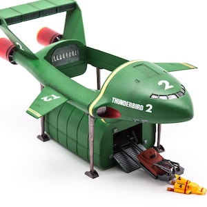 Puede incluir: Un modelo de avión Thunderbird 2 verde con el número 2 en las alas y el fuselaje, escapes de motor rojos, una cápsula de carga desmontable, una rampa, un vehículo marrón pequeño y una nave amarilla tipo submarino en el suelo.