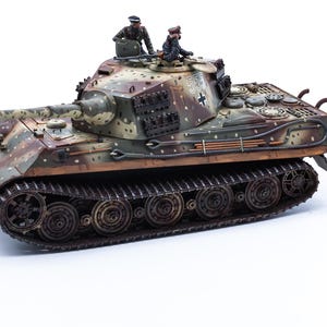 Op de afbeelding: Een schaalmodel van een Duitse King Tiger-tank met een camouflagepatroon van groene, bruine en beige vlekken. De tank heeft een lang kanon, donkere rupsbanden en twee miniatuurfiguren in het open luik van de koepel met het nummer 551.