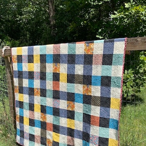 Puede incluir: Un edredón de retazos colorido con un patrón de cuadrícula, exhibido al aire libre. El edredón presenta cuadrados en varios colores, incluyendo negro, azul, amarillo y naranja. El edredón tiene un borde rosa y está colgado en una valla de madera.