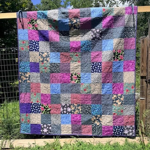 Op de afbeelding: Een patchwork quilt bestaande uit een raster van vierkante stoffen blokken in diverse tinten blauw, paars, roze en beige. De quilt toont een mix van effen kleuren en verschillende bloemen-, paisley- en stippenpatronen met zichtbare stiksels.