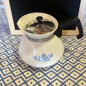 May include: White ceramic teapot with a black handle and lid. The teapot has a blue floral design and a silver lid. The handle is black and the rim has a decorative silver band. The teapot is on a blue and white patterned surface.