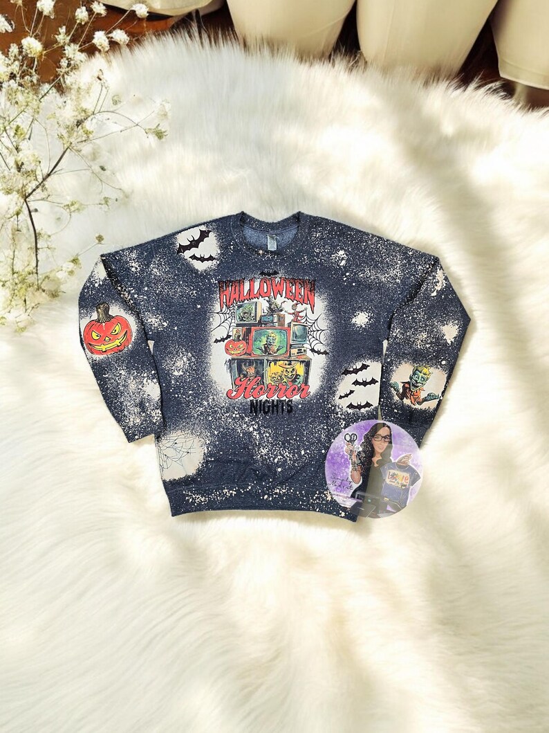 May include: A blue sweatshirt with a bleach-splattered design featuring a vintage television set with a Halloween theme. The text "Halloween Horror Nights" is printed on the sweatshirt.