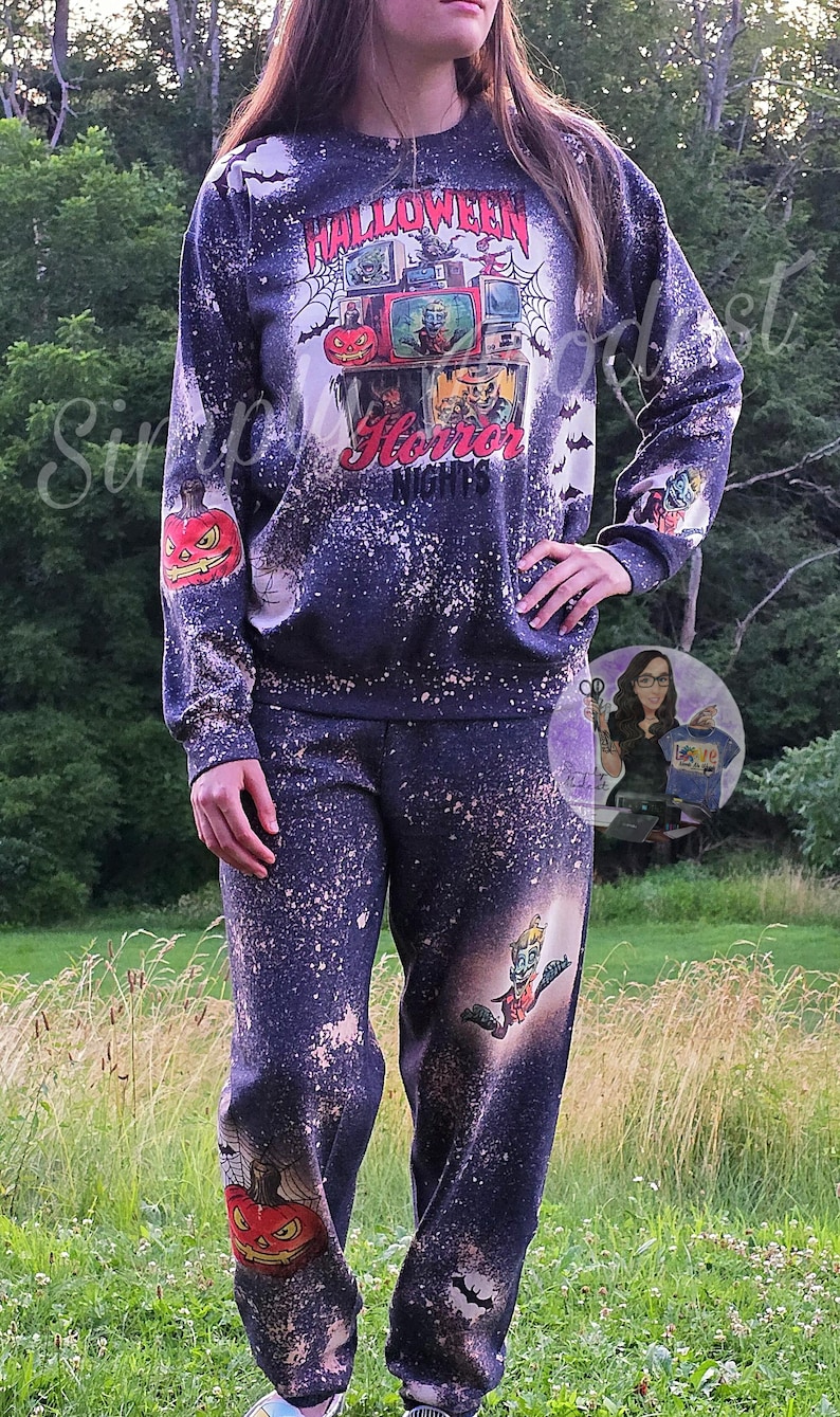 May include: A black and white bleached sweatshirt and sweatpants set with a Halloween horror nights theme. The sweatshirt features a graphic of a spiderweb with televisions and pumpkins. The sweatpants feature a graphic of a pumpkin and a monster.