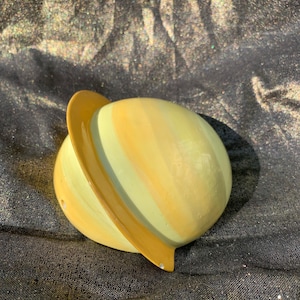 May include: A yellow and light green ceramic planet with a brown ring around the middle. The planet is sitting on a dark gray fabric surface.