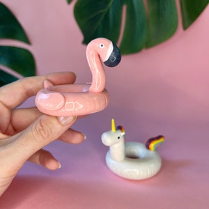 May include: Two ceramic figurines of inflatable pool floats. One is a pink flamingo and the other is a white unicorn with a rainbow tail.