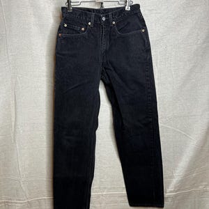 May include: Black denim jeans with a classic five-pocket design. The jeans are hung on a metal hanger. The jeans have a button and zipper closure, and rivets at the pockets. The denim appears to be a dark wash.