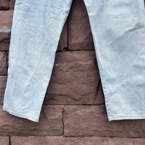 May include: Light blue denim jeans. The jeans are made of a light-wash denim material. The legs of the jeans are visible, with a slight fade and a relaxed fit. The background is a textured, reddish-brown brick wall.