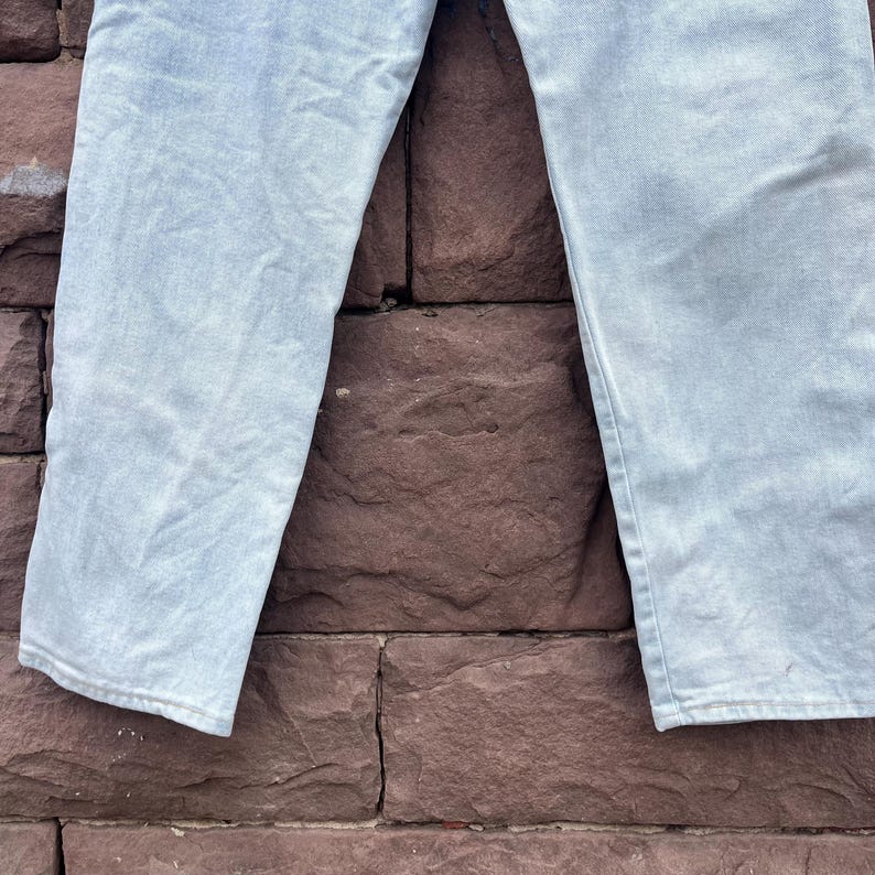 May include: Light blue denim jeans. The jeans are made of a light-wash denim material. The legs of the jeans are visible, with a slight fade and a relaxed fit. The background is a textured, reddish-brown brick wall.