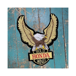 May include: Embroidered patch featuring a majestic eagle with outstretched wings. The eagle has a white head, brown body, and gold wings. Below the eagle is a banner with the words "GOLD HONDA WING" in gold lettering.