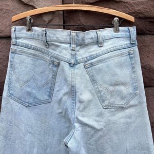 May include: Light blue denim jeans with the Wrangler logo on the back pockets. The jeans are hanging on a wooden hanger. The jeans have a classic five-pocket design and a straight leg cut.