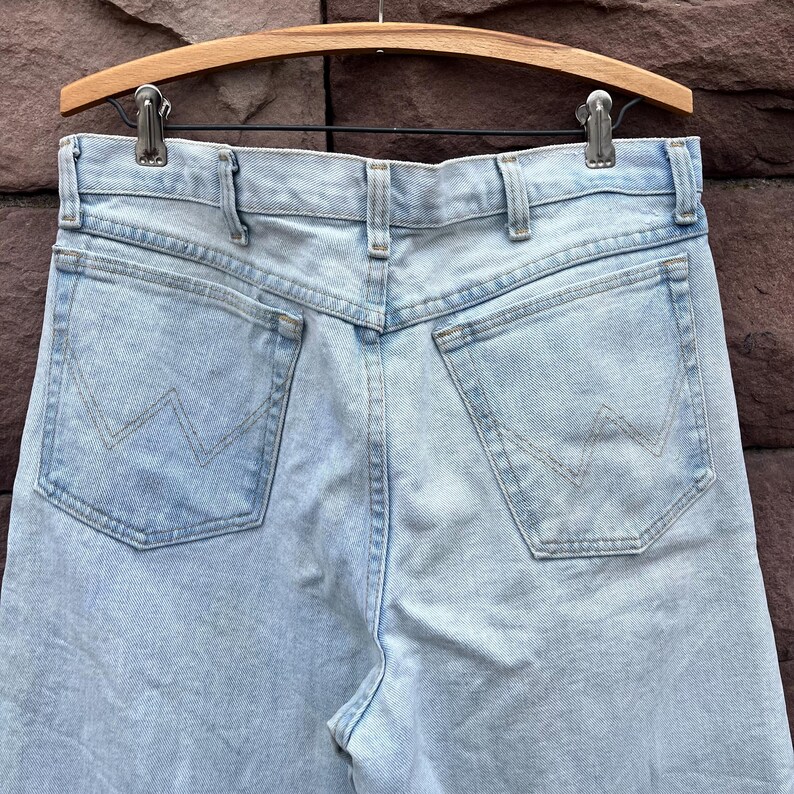 May include: Light blue denim jeans with the Wrangler logo on the back pockets. The jeans are hanging on a wooden hanger. The jeans have a classic five-pocket design and a straight leg cut.