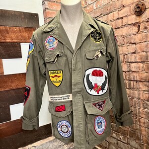 May include: Olive green military-style jacket with multiple patches. Patches include "Corsair Para Club", a parachute with wings, and "Galveston Skydivers". The jacket has multiple pockets and the text "CAUTION-CAUTION-CAUTION..." on the front.