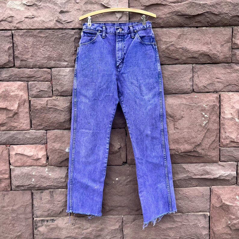 Puede incluir: Un par de vaqueros de mezclilla morados de cintura alta vintage colgados de una percha de madera. Los vaqueros tienen un dise&ntilde;o cl&aacute;sico de cinco bolsillos, cierre de bot&oacute;n y cremallera, y un dobladillo sin rematar deshilachado. Los vaqueros se exhiben contra una pared de ladrillos texturizados.