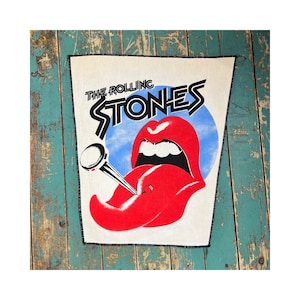 May include: A rectangular patch featuring the Rolling Stones logo. The design includes the band's name in black, a red tongue and lips graphic, and a blue circle. The patch is bordered with black stitching and is set against a white background.