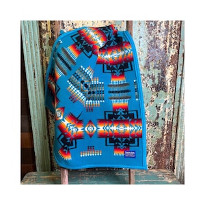 May include: A bright turquoise blanket featuring a repeating geometric pattern in shades of red, orange, yellow, white, and black. The fabric has a soft texture and a small rectangular blue label with white text in the bottom right corner.