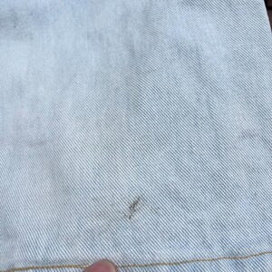 May include: Close-up of light blue denim fabric. The denim has a diagonal weave pattern and a few small, dark spots. The bottom edge of the fabric is visible, with a tan-colored seam.