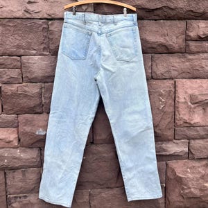 May include: Light blue denim jeans hanging on a wooden hanger. The jeans have a classic five-pocket design and a straight leg fit. The back pockets feature a subtle embroidered detail. The jeans are made of a light-wash denim material.