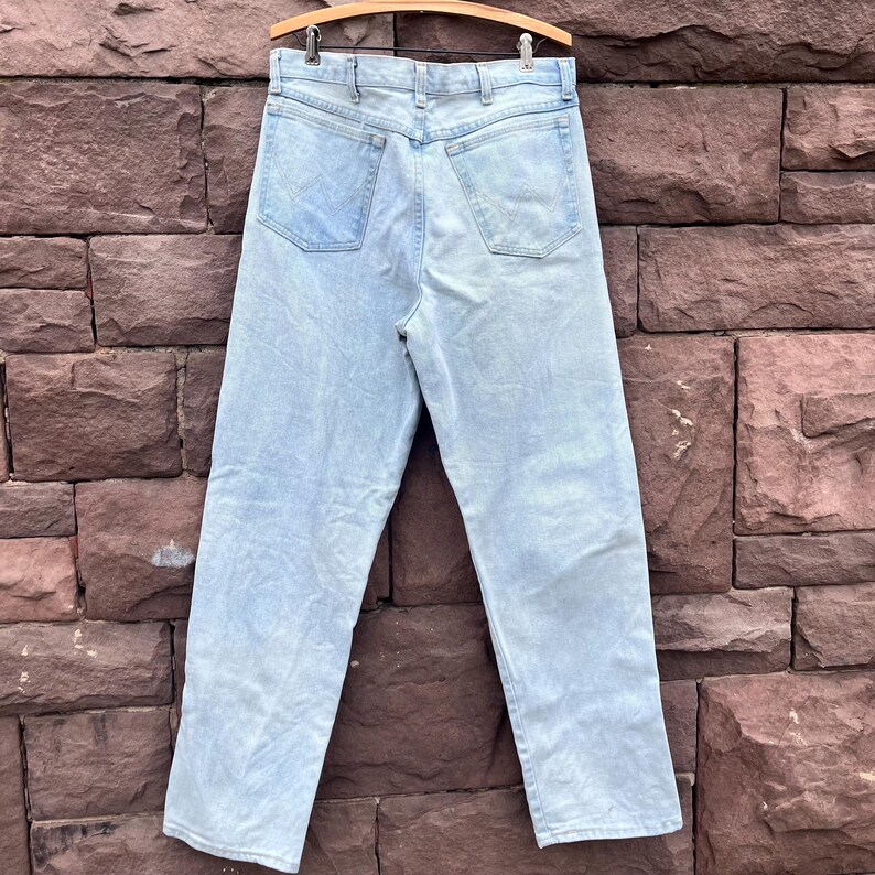 May include: Light blue denim jeans hanging on a wooden hanger. The jeans have a classic five-pocket design and a straight leg fit. The back pockets feature a subtle embroidered detail. The jeans are made of a light-wash denim material.