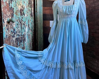 May include: A light blue vintage dress with long, puffy sleeves and a square neckline. The dress features white lace trim on the bodice, sleeves, and tiered skirt. The dress is displayed on a mannequin, showcasing its flowing, floor-length design.