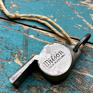 May include: A vintage metal whistle with the name "Wilson" and "MADE IN ENGLAND" stamped on the side. The whistle is attached to a beige rope. The background is a distressed, turquoise-painted wooden surface.