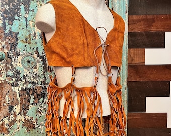May include: A tan suede vest with a V-neck and fringe detailing. The vest has a tie closure at the neckline and long fringe hanging from the bottom. The vest is displayed on a mannequin.