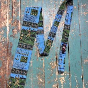 May include: A vintage golf-themed necktie with a blue background and green and yellow accents. The tie features illustrations of golf clubs, golfers, and text. The tie is made of a patterned fabric and is displayed on a weathered wooden surface.