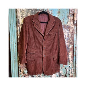 May include: A vintage brown corduroy blazer with a classic notched lapel, three-button closure, and two front pockets. The jacket is hanging on a black hanger against a distressed, turquoise and brown background.