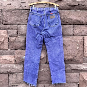 Puede incluir: Vaqueros de mezclilla azules vintage con un dise&ntilde;o cl&aacute;sico de cinco bolsillos, con el logotipo de Wrangler en los bolsillos traseros y un parche de cuero. Los vaqueros se exhiben en una percha de madera sobre un fondo de pared de ladrillo. Los vaqueros tienen una pernera recta y un dobladillo deshilachado.