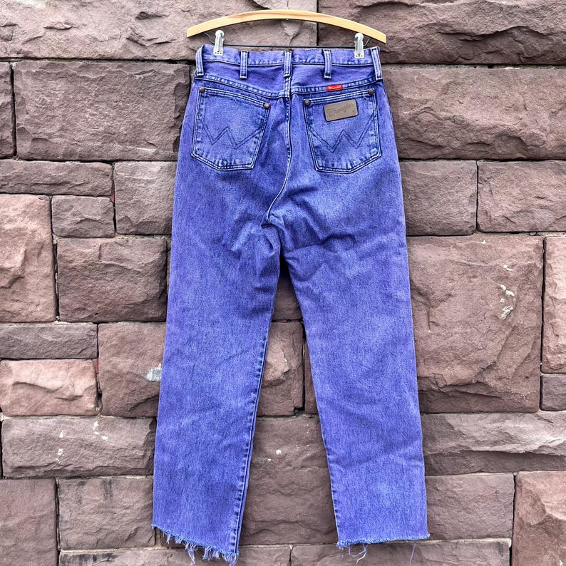 Puede incluir: Vaqueros de mezclilla azules vintage con un dise&ntilde;o cl&aacute;sico de cinco bolsillos, con el logotipo de Wrangler en los bolsillos traseros y un parche de cuero. Los vaqueros se exhiben en una percha de madera sobre un fondo de pared de ladrillo. Los vaqueros tienen una pernera recta y un dobladillo deshilachado.
