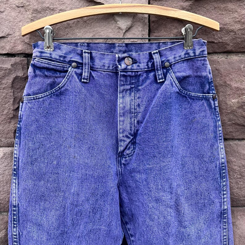 Puede incluir: Primer plano de unos vaqueros azules vintage colgados de una percha de madera. Los vaqueros tienen un aspecto descolorido, lavado al &aacute;cido, con cierre de bot&oacute;n y cremallera. Los vaqueros son de mezclilla y tienen bolsillos.