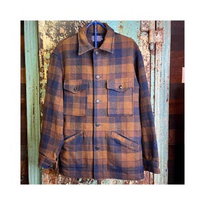 May include: A long-sleeve button-up jacket featuring a brown and navy blue plaid pattern. The garment includes a pointed collar, two chest pockets with buttoned flaps, and two lower slanted pockets. A Pendleton brand label is visible at the neck.