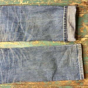 Pode incluir: Close-up da parte inferior de um par de jeans azul claro. Os jeans t&ecirc;m uma apar&ecirc;ncia desbotada com uma bainha dobrada na parte inferior. O denim mostra desgaste, com vincos vis&iacute;veis e manchas mais claras.