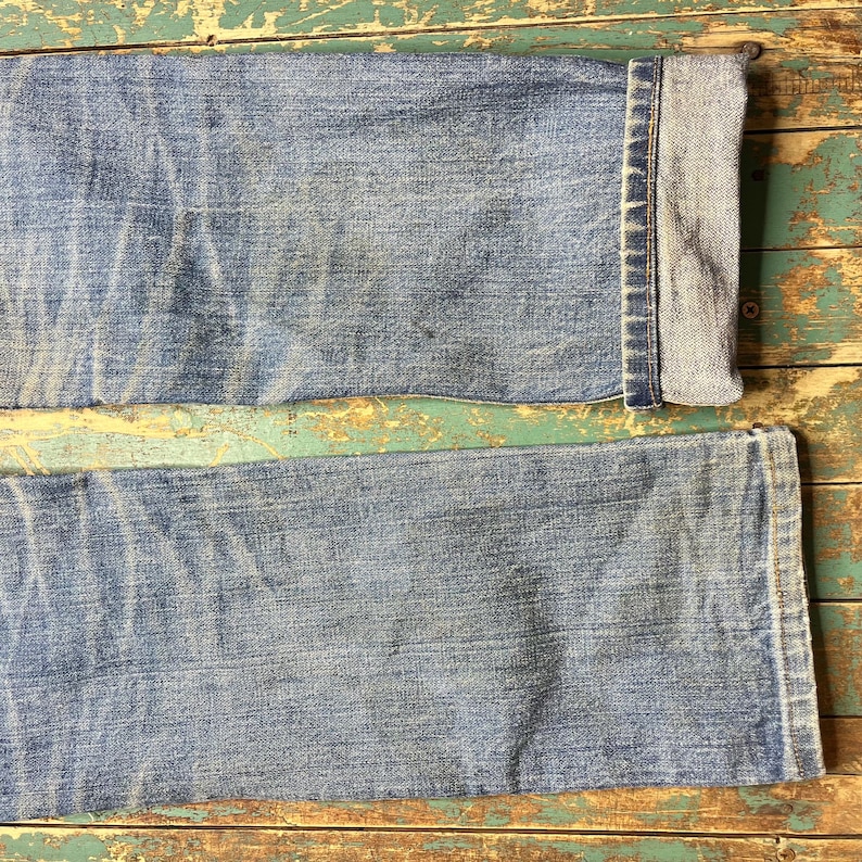 Pode incluir: Close-up da parte inferior de um par de jeans azul claro. Os jeans t&ecirc;m uma apar&ecirc;ncia desbotada com uma bainha dobrada na parte inferior. O denim mostra desgaste, com vincos vis&iacute;veis e manchas mais claras.